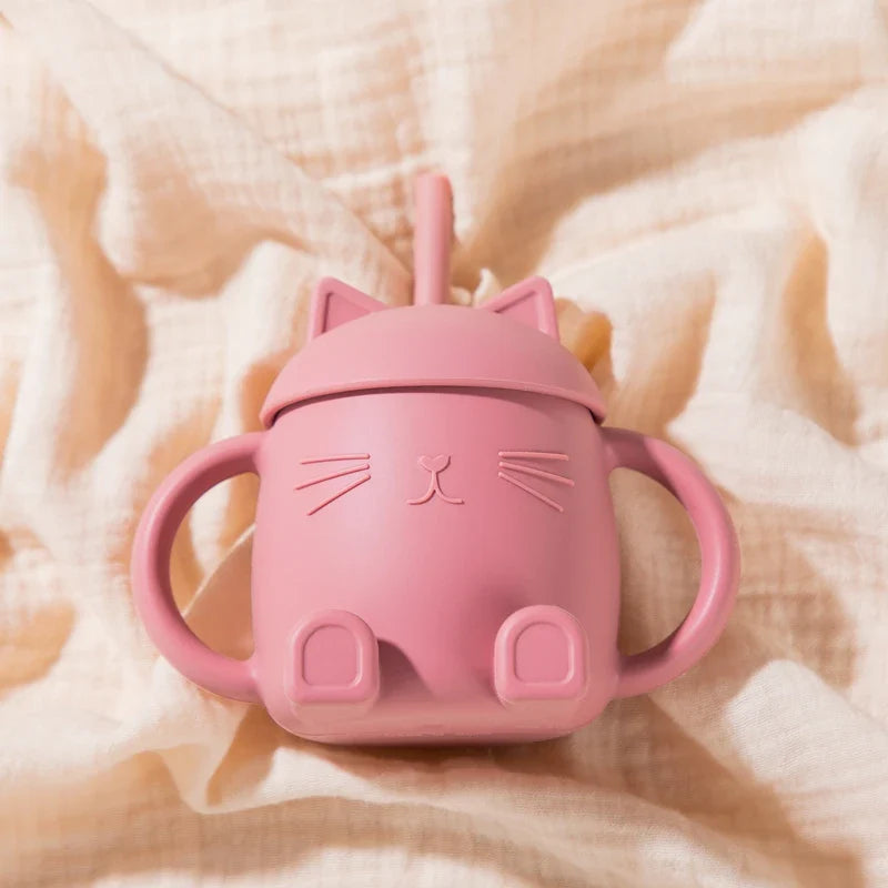 Der Hello World Trinkbecher Katze sitzend Pink [13x11cm] ist ein rosafarbener Becher in Form einer Katze mit zwei Henkeln, Katzenohren auf dem Deckel, Schnurrhaaren und Nasendetails sowie kleinen Füßen am Boden, der auf einem weichen beigen Stoff ruht.