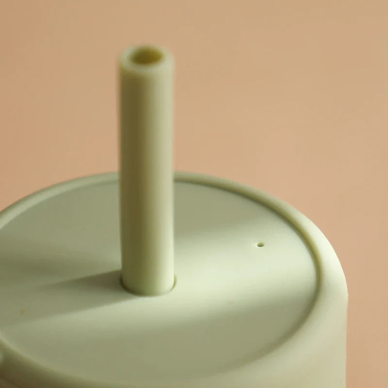 Eine Nahaufnahme des Hello World Trinkbechers mit Motiv Grün [6x12,5cm] zeigt den blassgrünen, BPA-freien Silikondeckel und den passenden Strohhalm vor einem zartbeigen Hintergrund.