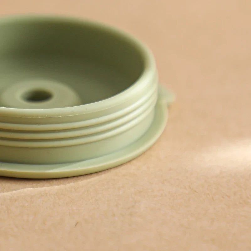 Eine Nahaufnahme eines hellgrünen, runden Kunststoffdeckels mit Gewinde, der auf einer beigen Oberfläche ruht - entworfen für den Hello World Trinkbecher mit Motiv Grün [6x12,5cm].