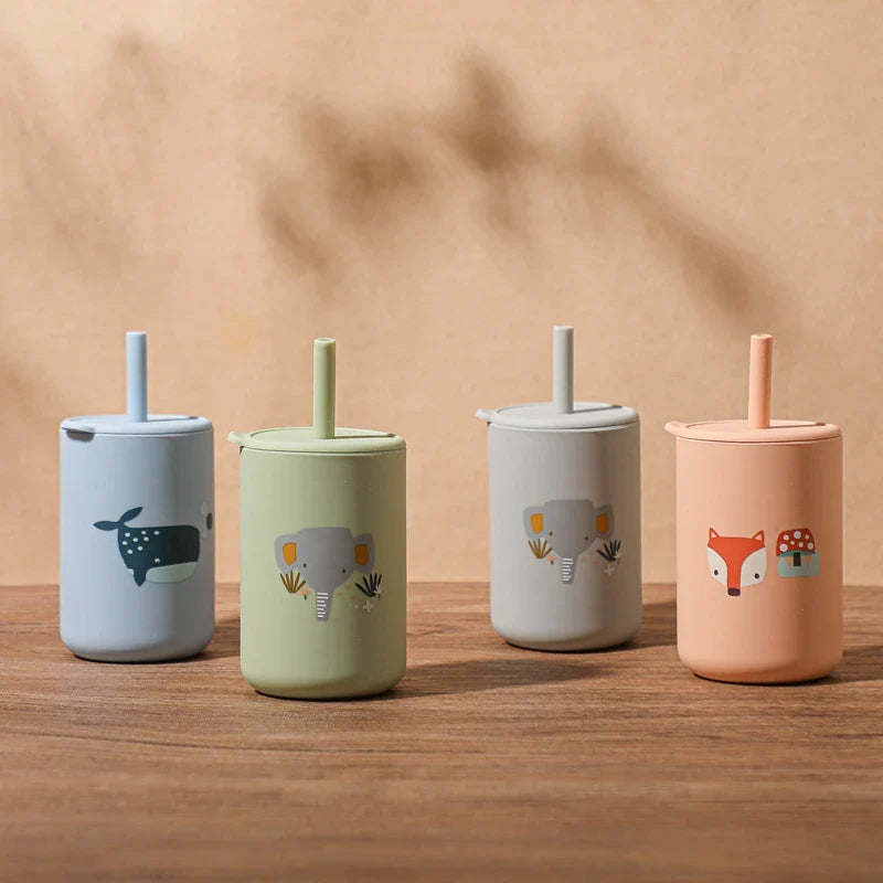 Vier Hello World Trinkbecher mit Motiv Orange [6x12,5cm], aus BPA-freiem Silikon, mit Deckel und Strohhalm im Fuchs-Design und weiteren Tiermotiven (Wal, Elefant), stehen auf Holz vor beigem Hintergrund.