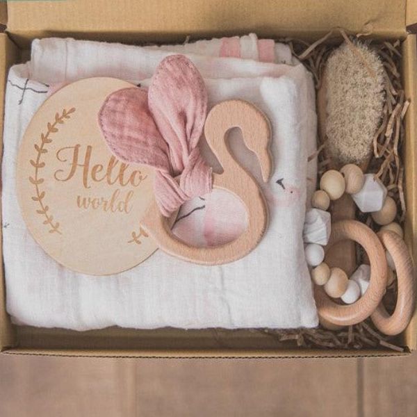 Eine Person hält das Victoria Baby Geschenk zur Geburt von Hello World in der Hand: eine Schachtel mit einem hölzernen "Hello World"-Schild, einem Holz-Silikon-Beißring mit rosa Schwanenohren, einem Holz-Greifling, einem Naturschwamm und einem gefalteten weißen Tuch - ideal als Geschenk zur Geburt.