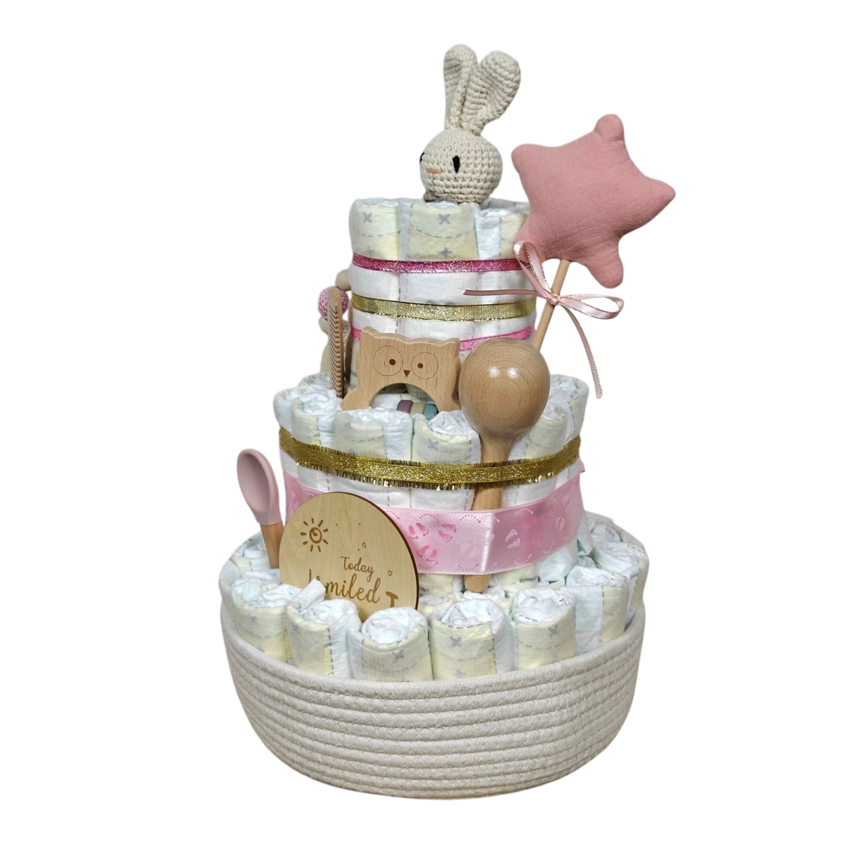 Die Hello World Windeltorte Hase [25x42cm] besteht aus drei Etagen mit rosa und goldenen Bändern, einem gehäkelten Hasen, einem Sternenstab, Holzspielzeug und Babyartikeln in einem geflochtenen Korb - ein ideales nachhaltiges Babygeschenk und Geschenk zur Geburt.