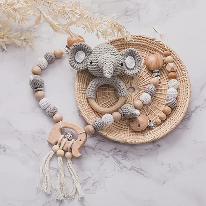 Handgefertigtes Babyzubehör mit einem gehäkelten Elefanten-Beißring, Hello World Kinderwagenkette Elefant [20x32cm], und perlenbesetzten Schnullerclips in Natur- und Grautönen, die alle auf einem gewebten runden Tablett angeordnet sind.