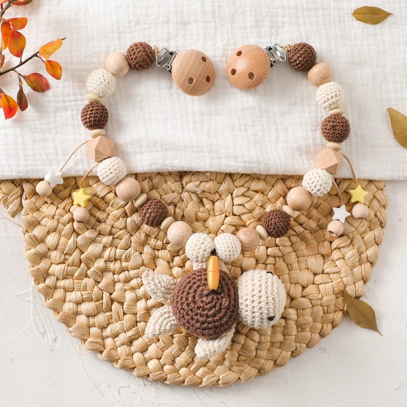 Die Hello World Kinderwagenkette Schildkröte [30x31cm] besteht aus hölzernen und gehäkelten Perlen in neutralen Tönen sowie einer gehäkelten Schildkröte in der Mitte - ideal für entspannte Reisen. Abgebildet auf einer gewebten Matte, die mit Herbstblättern verziert ist.