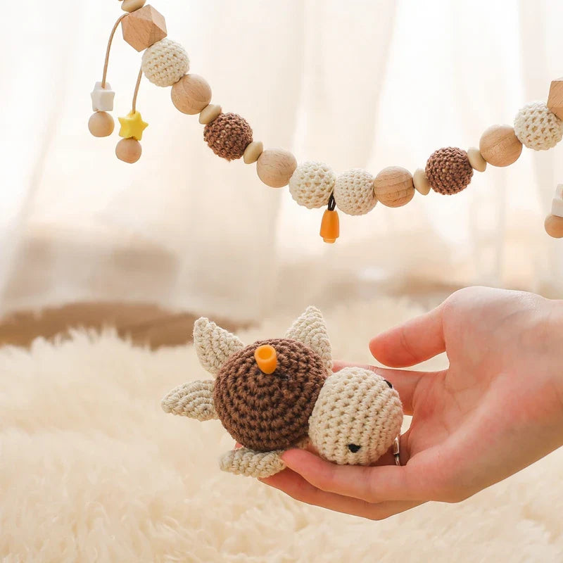 Eine Hand hält eine kleine beige und braune gehäkelte Schildkröte, während die Hello World Kinderwagenkette Schildkröte [30x31cm] mit hölzernen und gehäkelten Perlen oben auf einer flauschigen Oberfläche hängt - perfekt für entspannte Reisen.