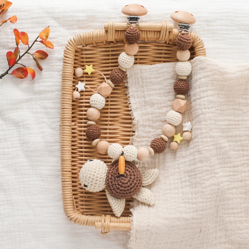 Ein geflochtener Korb enthält ein beiges Tuch und die Hello World Kinderwagenkette Schildkröte [30x31cm] mit Holz-, Häkel- und Sternperlen sowie einer Schildkröte - ideal für entspanntes Reisen. Die Herbstblätter sorgen für zusätzliche Gemütlichkeit.