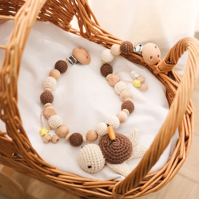 Die Hello World Kinderwagenkette Schildkröte [30x31cm] besteht aus Holz- und Häkelperlen in Natur-, Braun- und Gelbtönen mit einem Schildkrötenspielzeug - ein bezauberndes Accessoire für entspannte Reisen, abgebildet auf einem weißen Tuch in einer Korbwiege.