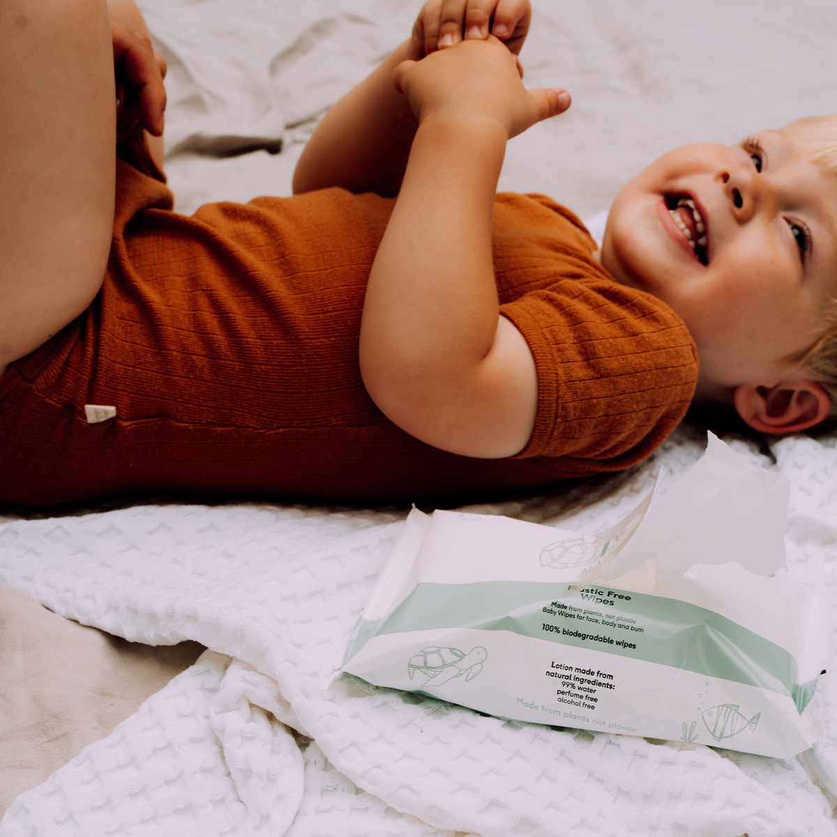 Naïf plastic-free baby wipes next to a smiling baby on a soft blanket.