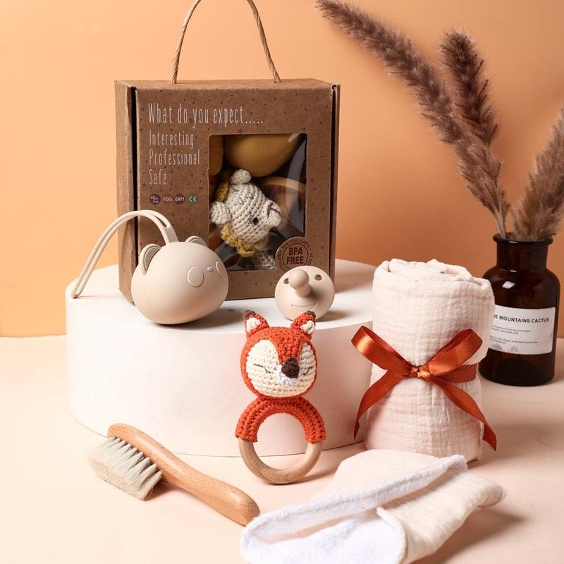 Das Rico Baby Geschenk zur Geburt von Hello World enthält eine Rasselfuchs-Rassel, eine Babybürste, ein Schnuller-Set, einen Beißring, ein aufgerolltes Tuch mit orangefarbenem Band und eine Pflanze in einer braunen Vase auf einem weißen Podest.
