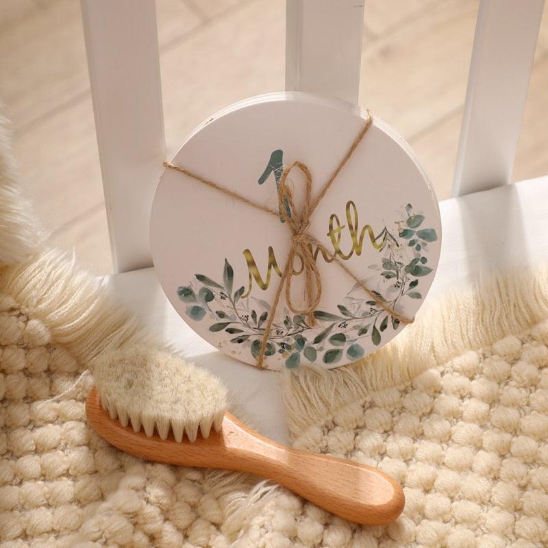 A round, floral milestone sign reading "1 Month" from Hello World's Rusty Baby Newborn Gift rests on a cream rug next to a baby hairbrush and white crib bars.