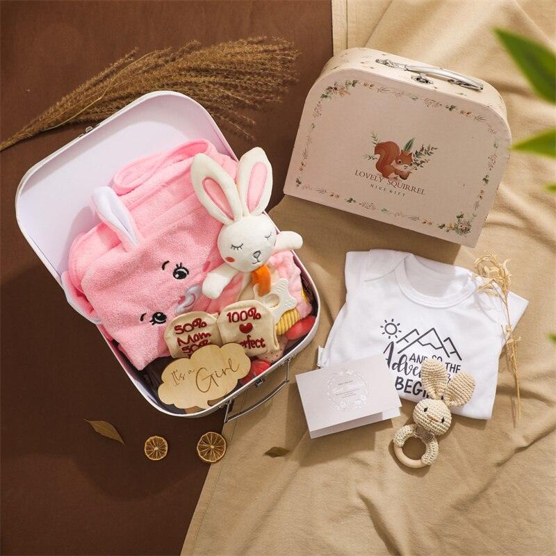 Das Selma Baby Geschenk zur Geburt von Hello World besteht aus einem rosa Häschenhandtuch, weißem Häschenplüsch, einer Holzrassel, Babykleidung, einem gehäkelten Häschen und einer geblümten Schachtel - ein ideales Babygeschenk auf einem gemütlichen braun-beigen Hintergrund.