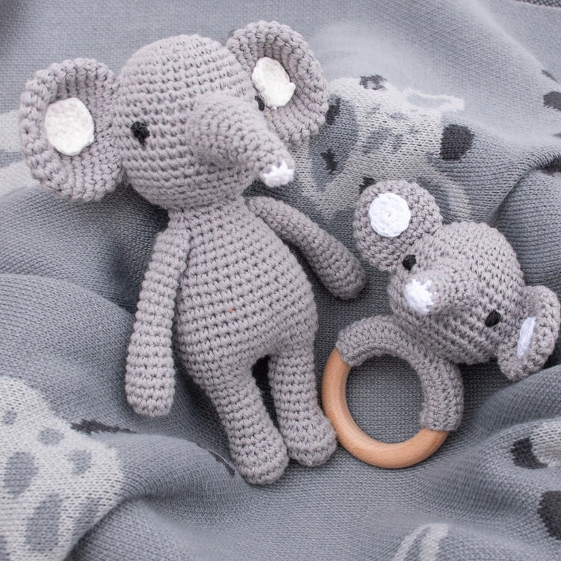 Auf einer weichen, grauen Decke liegen zwei Hello World Stricktier Elefant (18x12cm) - ein kuscheliger Plüschelefant und eine Elefantenrassel mit Holzringgriff - ideal für jedes Kuscheltier-Safari-Abenteuer.