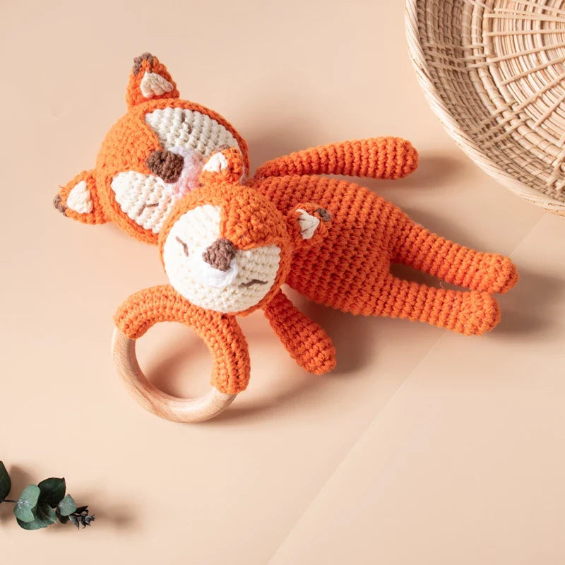 Due volpi arancioni lavorate a maglia all'uncinetto (18,5 x 11,5 cm) di Hello World, un tenero peluche e un giocattolo per la dentizione, giacciono su una superficie luminosa accanto a un cestino intrecciato e del verde.