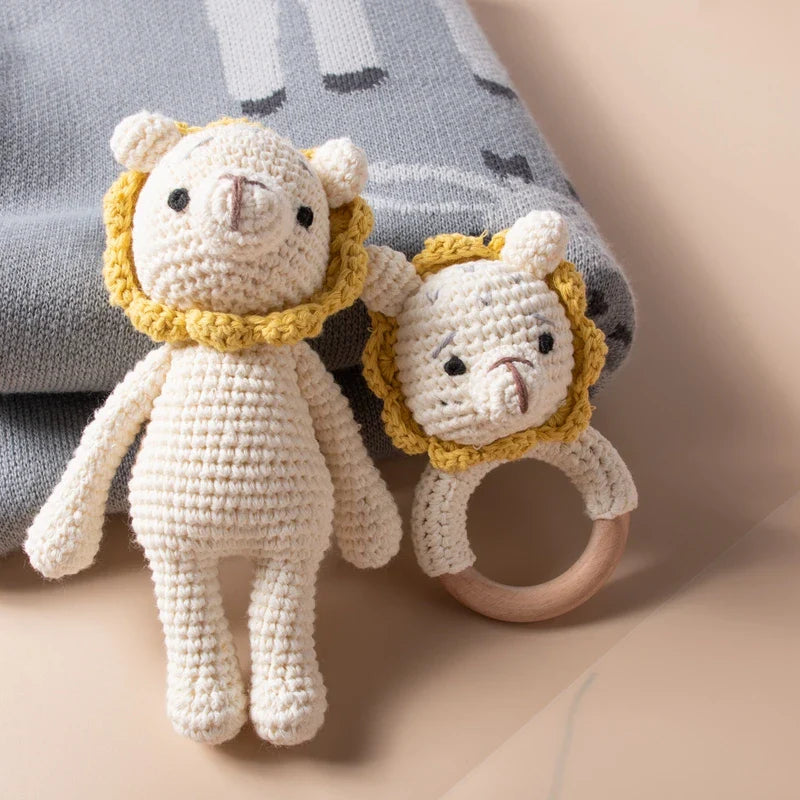 Due leoni lavorati a maglia Hello World (18,5x10 cm), un peluche e un sonaglio ad anello, entrambi con corpo color crema e criniera gialla, sono presentati su una morbida coperta grigia e bianca.