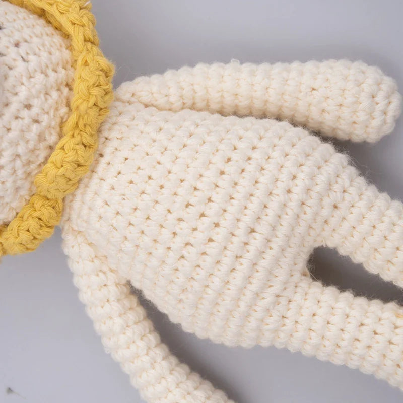 Primo piano del leone giocattolo lavorato a maglia Hello World (18,5x10 cm), un leone di peluche color crema con una criniera gialla, appoggiato su una superficie grigio chiaro. La foto mette in risalto il suo corpo strutturato e le sue braccia.