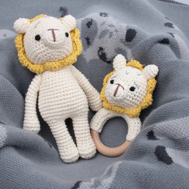 Due leoni fatti a mano a maglia Hello World (18,5x10 cm) - un peluche, un sonaglio con un anello di legno - giacciono su una coperta grigia con corpi color crema e criniere gialle.