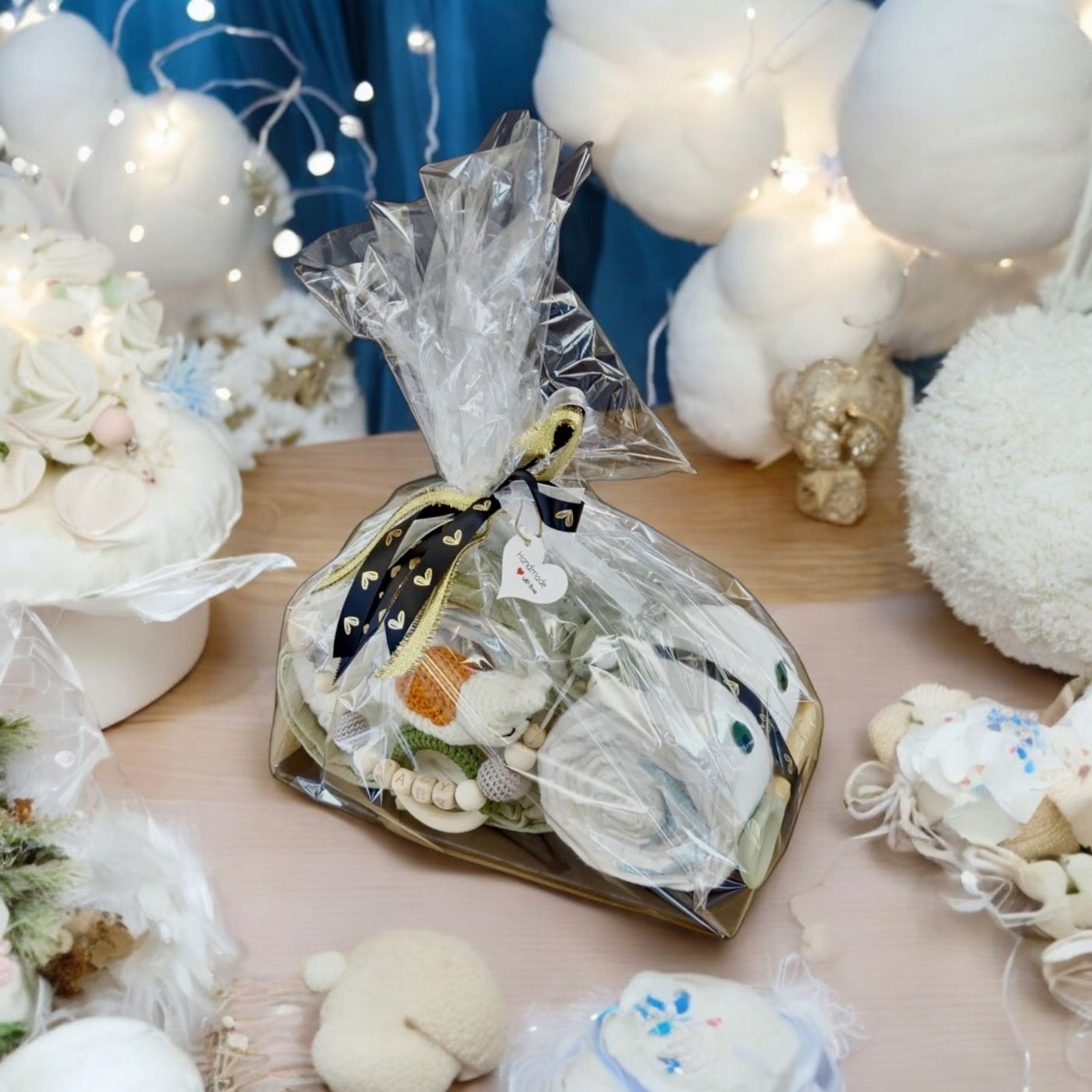 Auf einem Holztisch, umgeben von weißer Dekoration, Lichterketten und pastellfarbenen Gegenständen, liegt eine Hello World "Transparente Folie"-Geschenktüte, eingewickelt in durchsichtiges Plastik mit einem schwarzen Band und einem herzförmigen Anhänger.