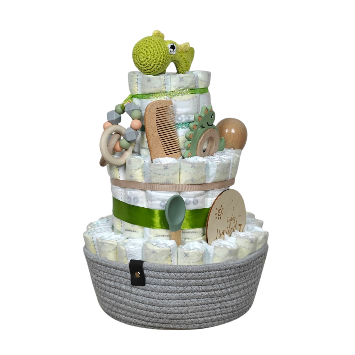 Die Windeltorte Dino [25x38cm] von Hello World ist dreistöckig, mit grünen Bändern, natürlichen Baby-Essentials, Holzspielzeug, Haarbürste und gehäkeltem grünem Hippo im grauen Korb - ein liebevolles Geschenk zur Geburt.